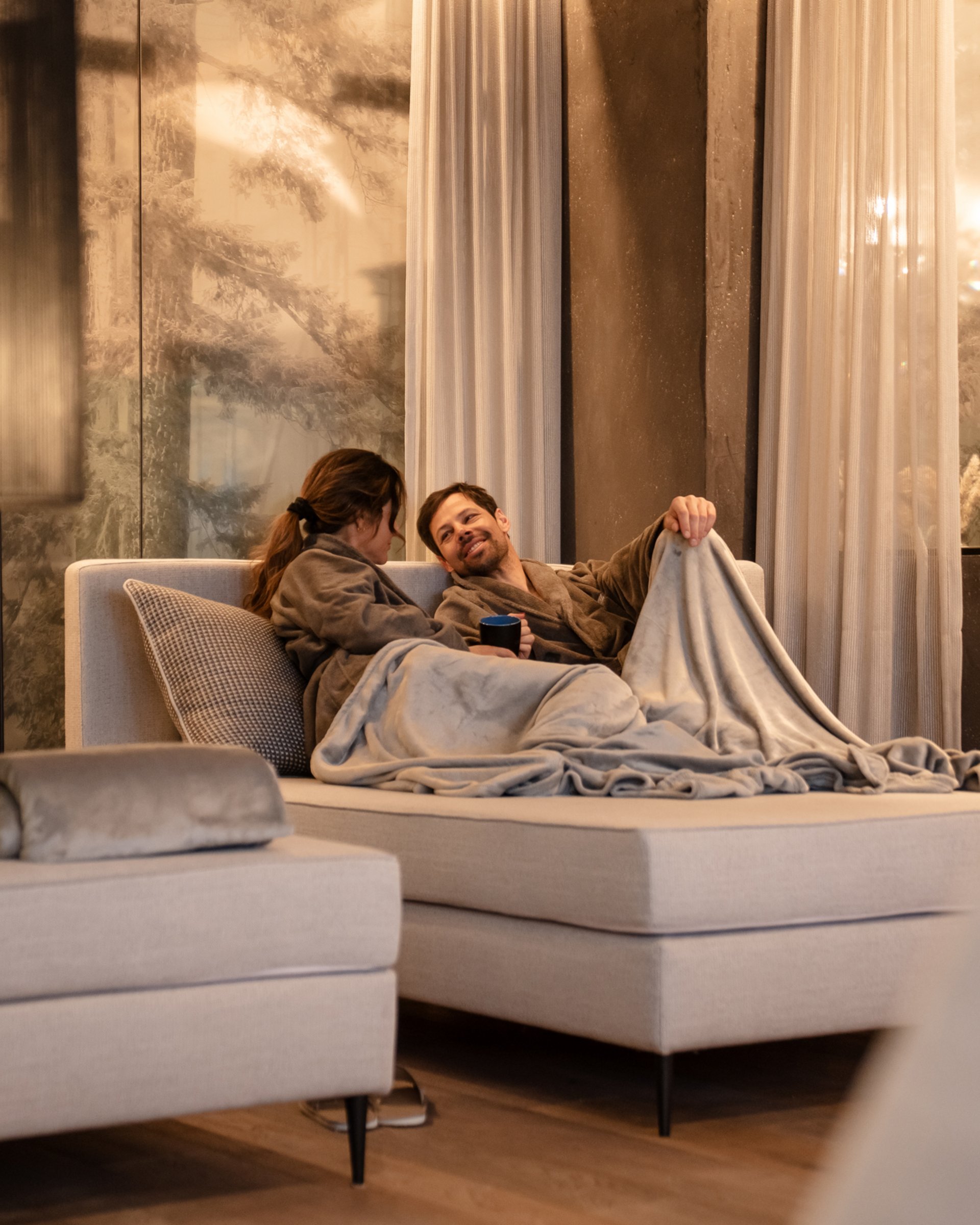 Wohlfühlurlaub im 5-Sterne-Hotel in Serfaus Paar in Bademänteln kuschelt sich auf Sofa in gemütlichem Raum mit warmem Licht