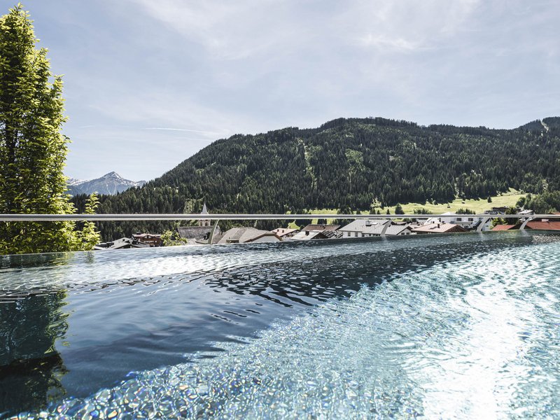 Wohlfühlurlaub im 5-Sterne-Hotel in Serfaus Infinity-Pool mit Aussicht auf grüne Berge und Dorf bei klarem Himmel
