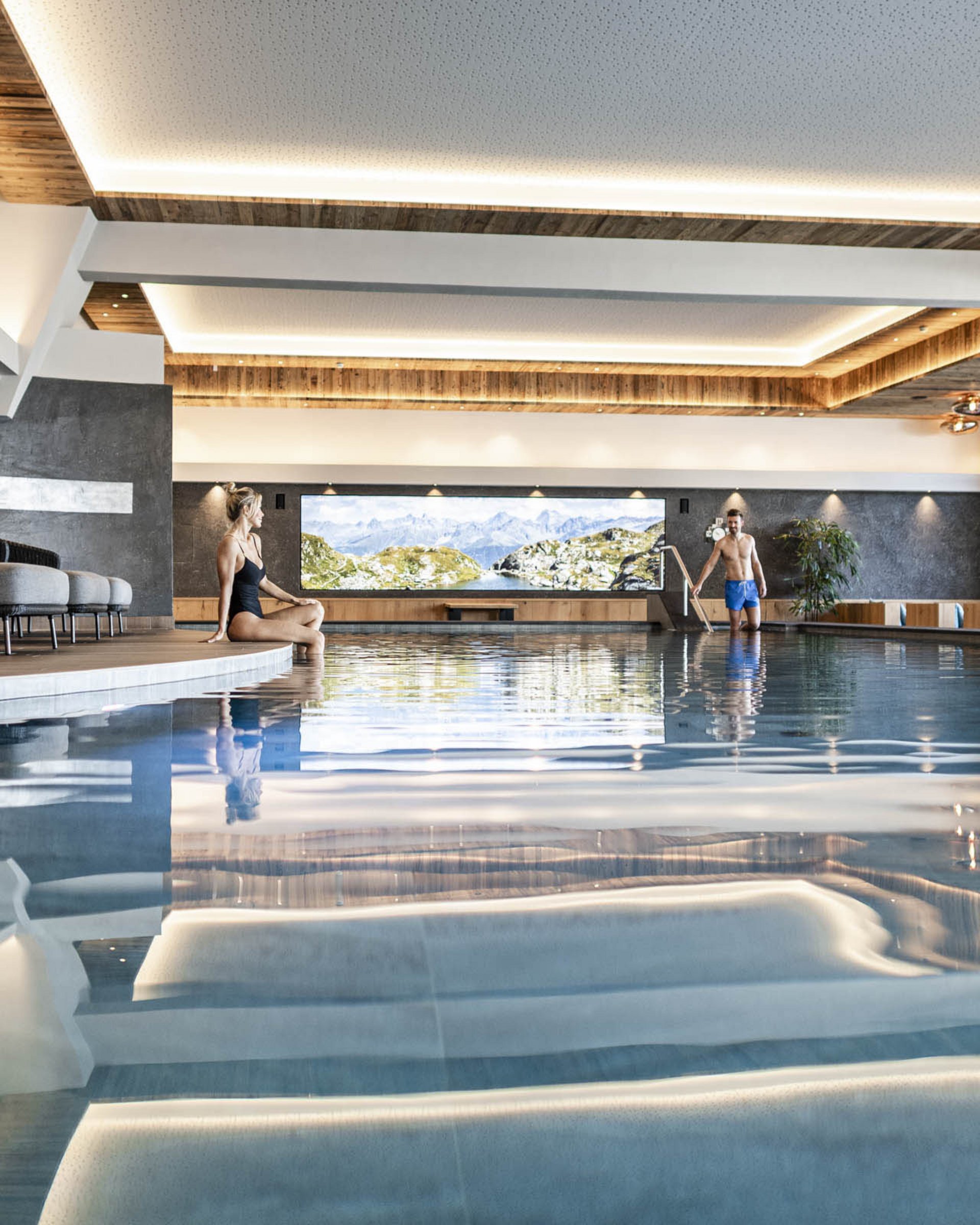 Wohlfühlurlaub im 5-Sterne-Hotel in Serfaus Innenraum mit Schwimmbad, Frau sitzt am Beckenrand, Mann steigt ins Wasser
