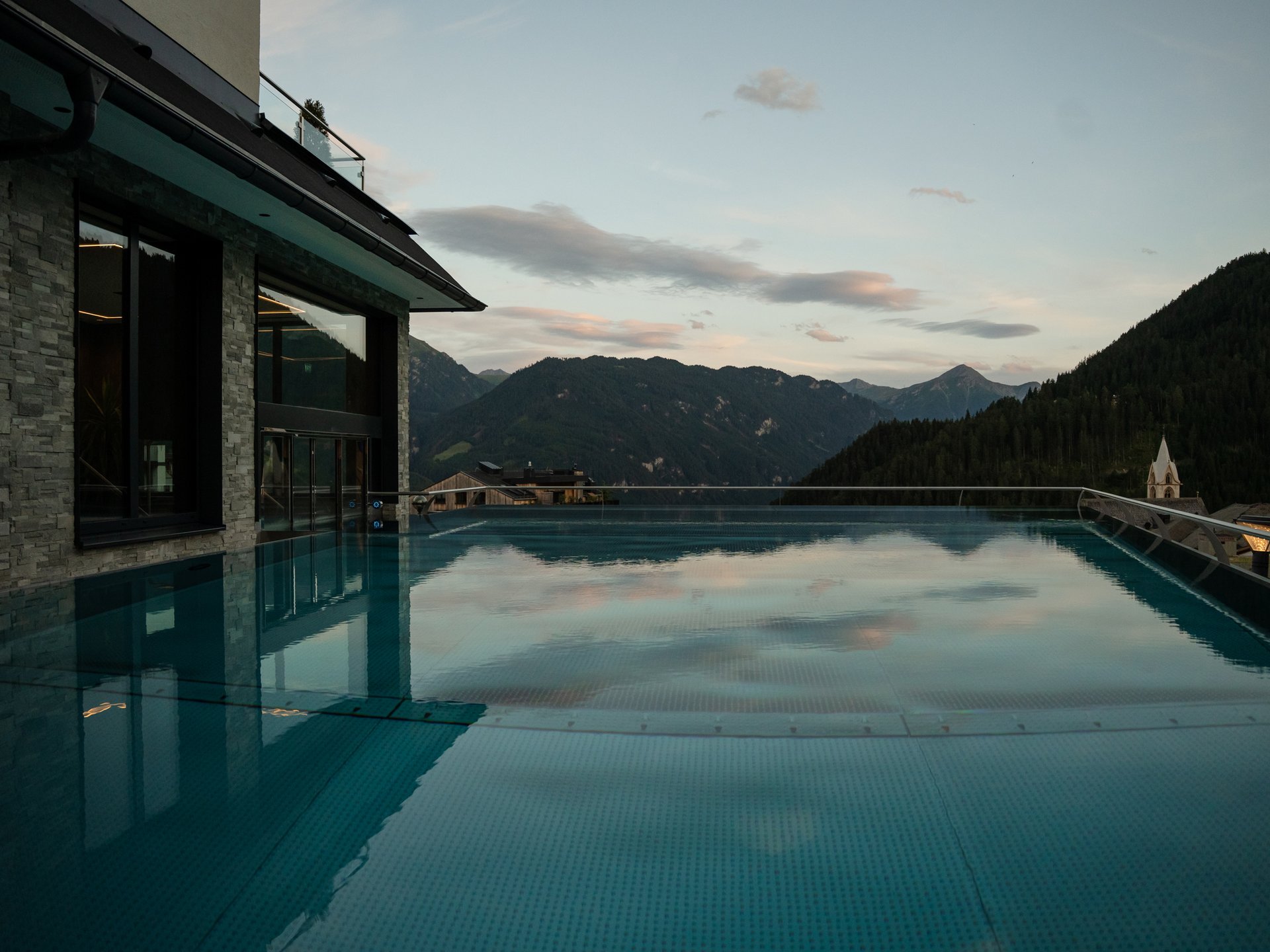 Wohlfühlurlaub im 5-Sterne-Hotel in Serfaus Infinity-Pool mit Bergblick und Abendhimmel neben modernem Gebäude