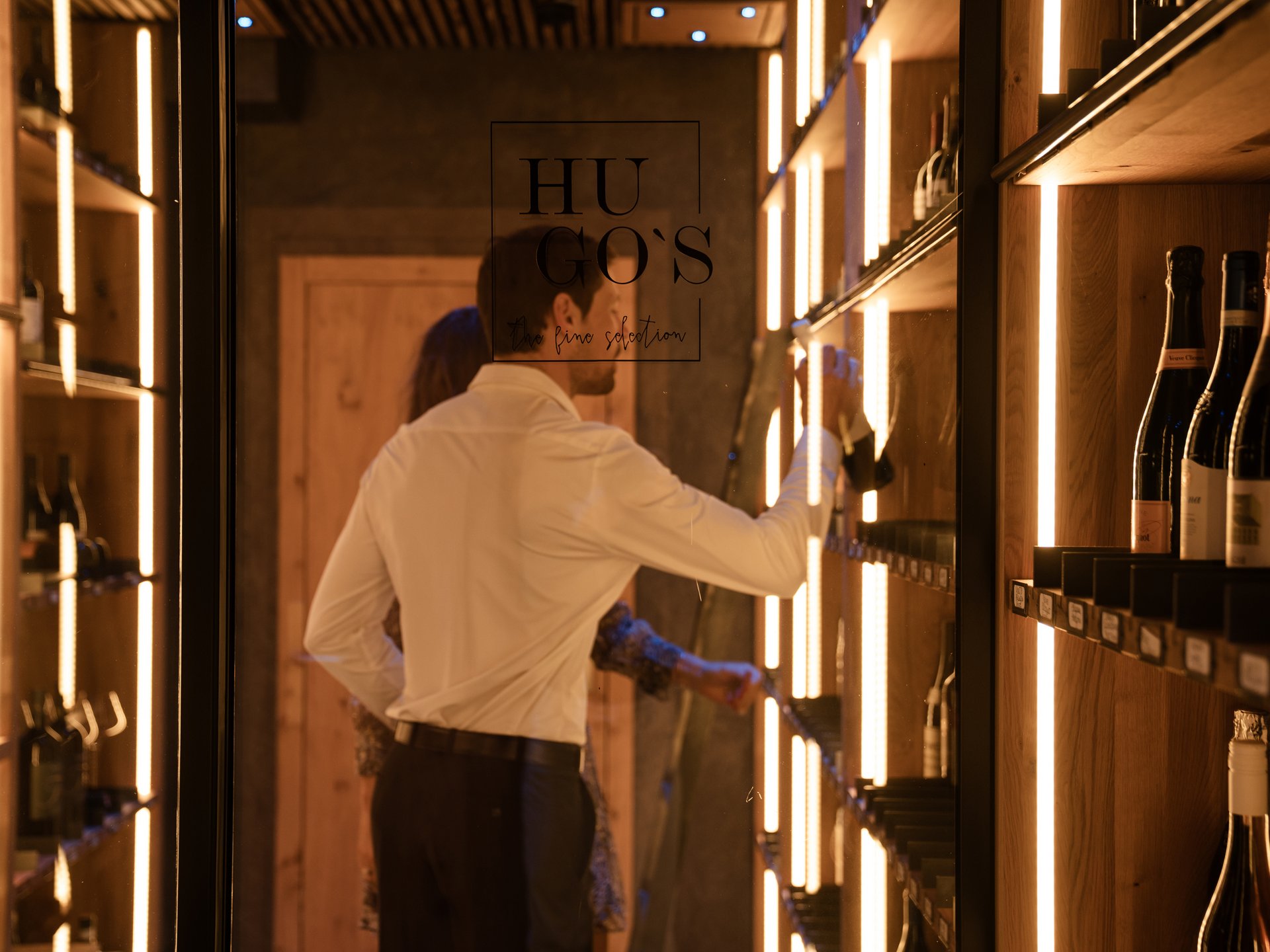 Finest wines and more: wine holiday in Austria at Cervosa Man selects wine bottle in stylish wooden wine cabinet with warm lighting