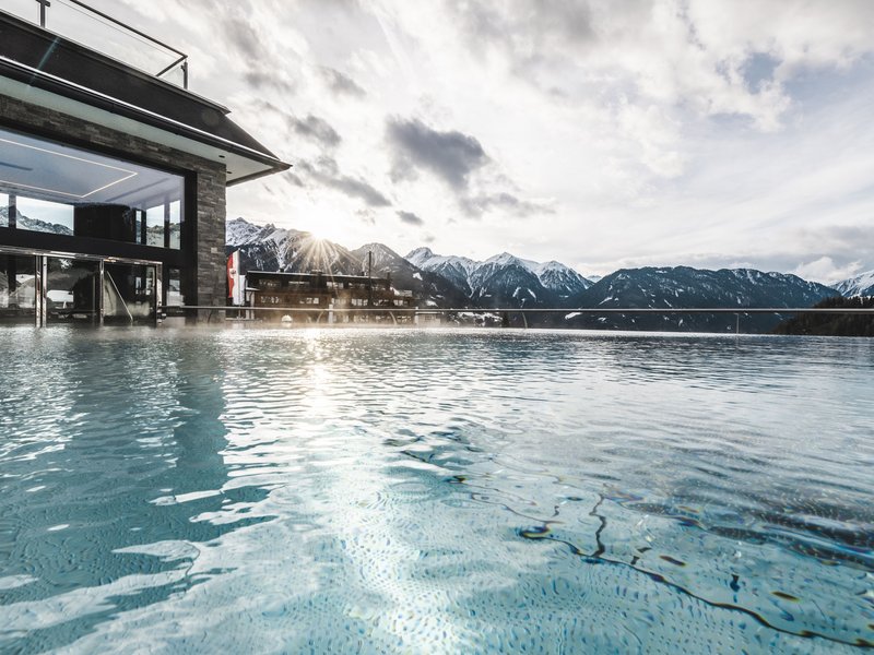 Wohlfühlurlaub im 5-Sterne-Hotel in Serfaus Außenpool mit Bergblick und Sonnenschein an einem bewölkten Tag